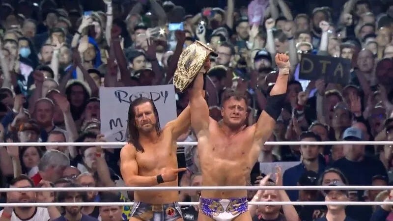 Adam Cole, long hair, beard and moustache, white guy, a little scrawny, holding up the arm and pointing at MJF, a beefier guy with curly hair.