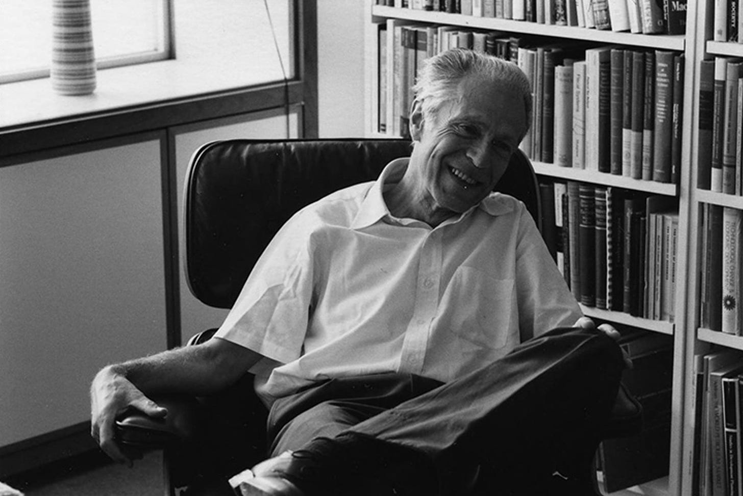 Vintage photo of an elderly man smiling, sitting in a chair in a room with a bookshelf in the background.