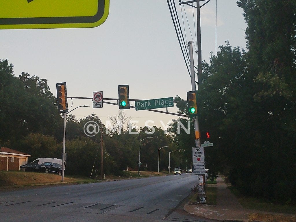A photo of a traffic light at the intersection of Forest Park boulevard and Park place in Fort worth, texas. The traffic light pole is leaning thanks to having been crashed into at its base by a reckless driver. The pole is leaning in such a way that it is straining some utility lines and appears to be in danger of falling.