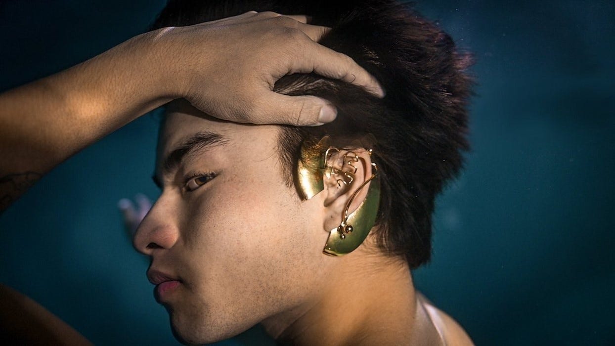 side view of a young man wearing hearing aid jewelry—a shiny brass c-shaped ear piece that wraps around the outer ear and has decorative wire covering the upper lobe
