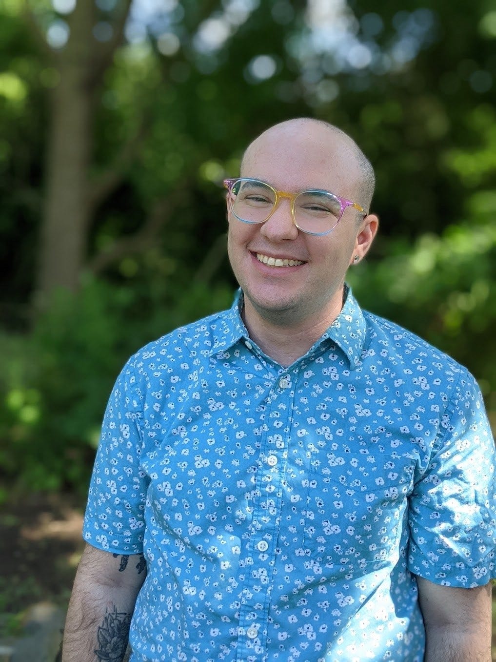 char hersh wears a blue print shirt and wears glasses. They are smiling and standing in front of a background of trees