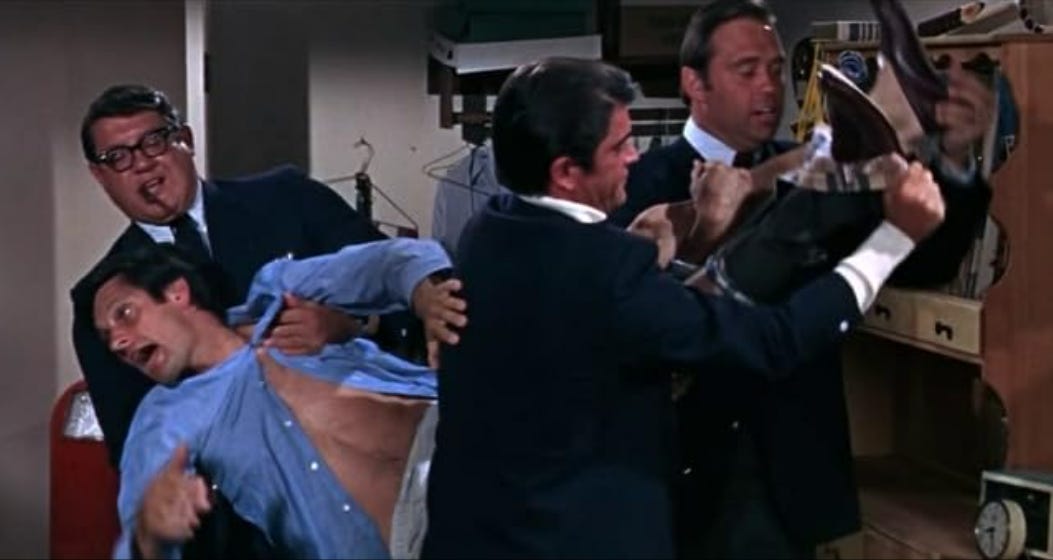 The actor Alan Alda, half-dressed, comically yells in surprise as three men in suits, including the athlete and actor Alex Karras wearing black horn-rimmed glasses, carry Alda from his bedroom.