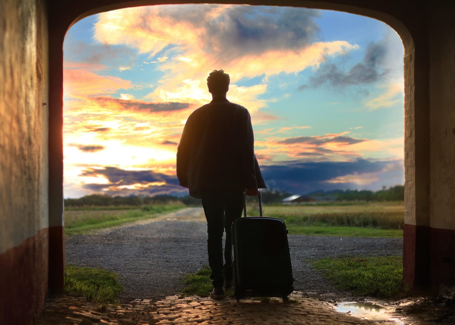 A man walks away from the camera into a sunset, wheeling a suitcase 