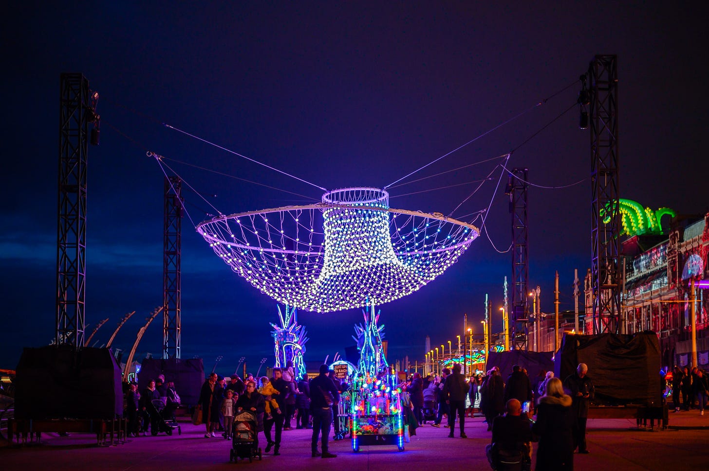 A cloud of LED lights hangs over a promenade at dusk as people walk underneath.