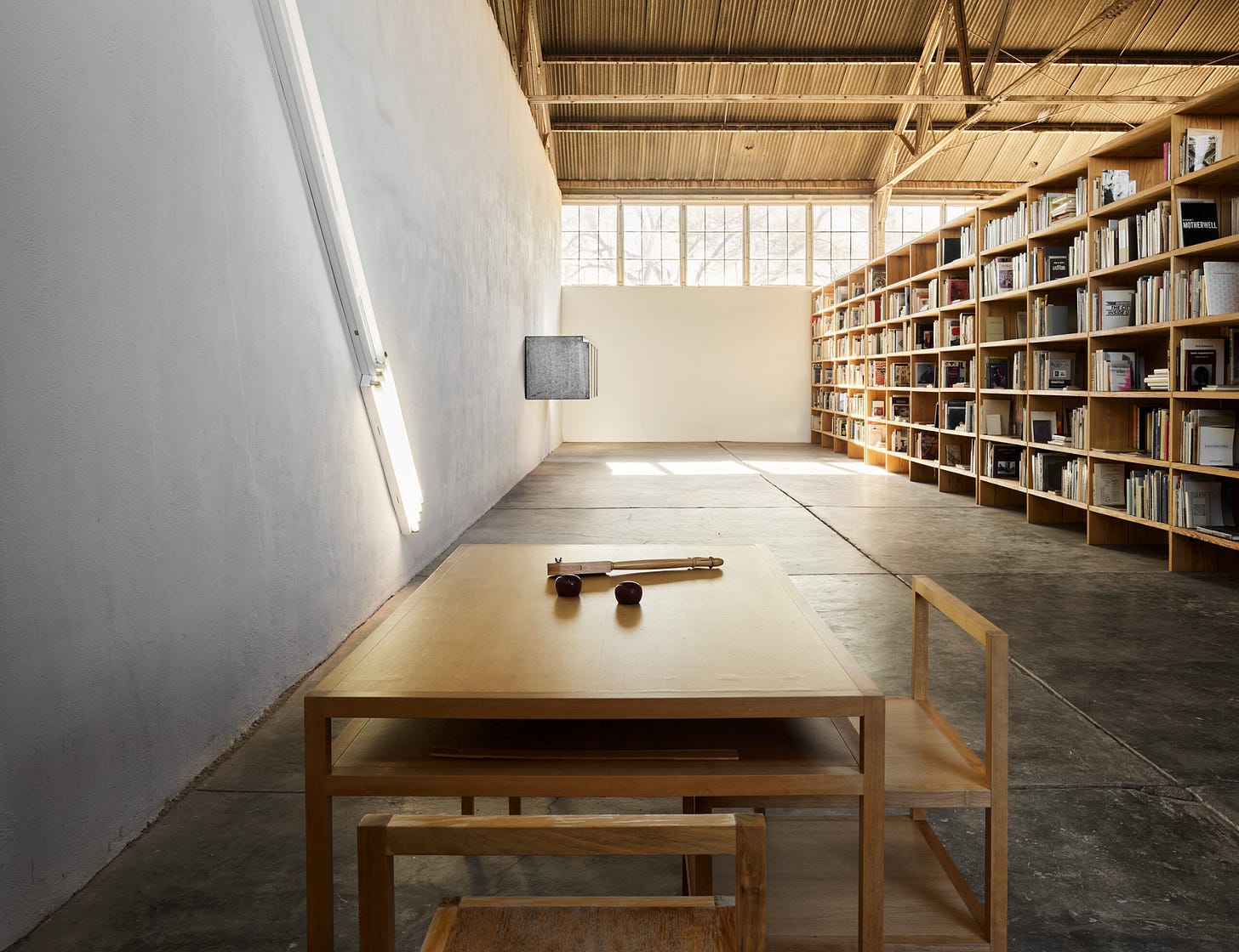A photograph of sculptor Donald Judd’s library at his Marfa, Texas home. The building housing the library is a former airplane hanger with high ceilings. A row of windows under the ceiling lets in a flood of light. The view is of one end of the library. To the left you can see a wall with sculptures by Dan Flavin and Judd. To the right is a very large and tall bookshelf with large art books on it. In the foreground is a table with two chairs and three ambiguous objects placed carefully on top. The feeling is light-filled and spacious. A photograph of sculptor Donald Judd’s library at his Marfa, Texas home. The building housing the library is a former airplane hanger with high ceilings. A row of windows under the ceiling lets in a flood of light. The view is of one end of the library. To the left you can see a wall with sculptures by Dan Flavin and Judd. To the right is a very large and tall bookshelf with large art books on it. In the foreground is a table with two chairs and three ambiguous objects placed carefully on top. The feeling is light-filled and spacious.
