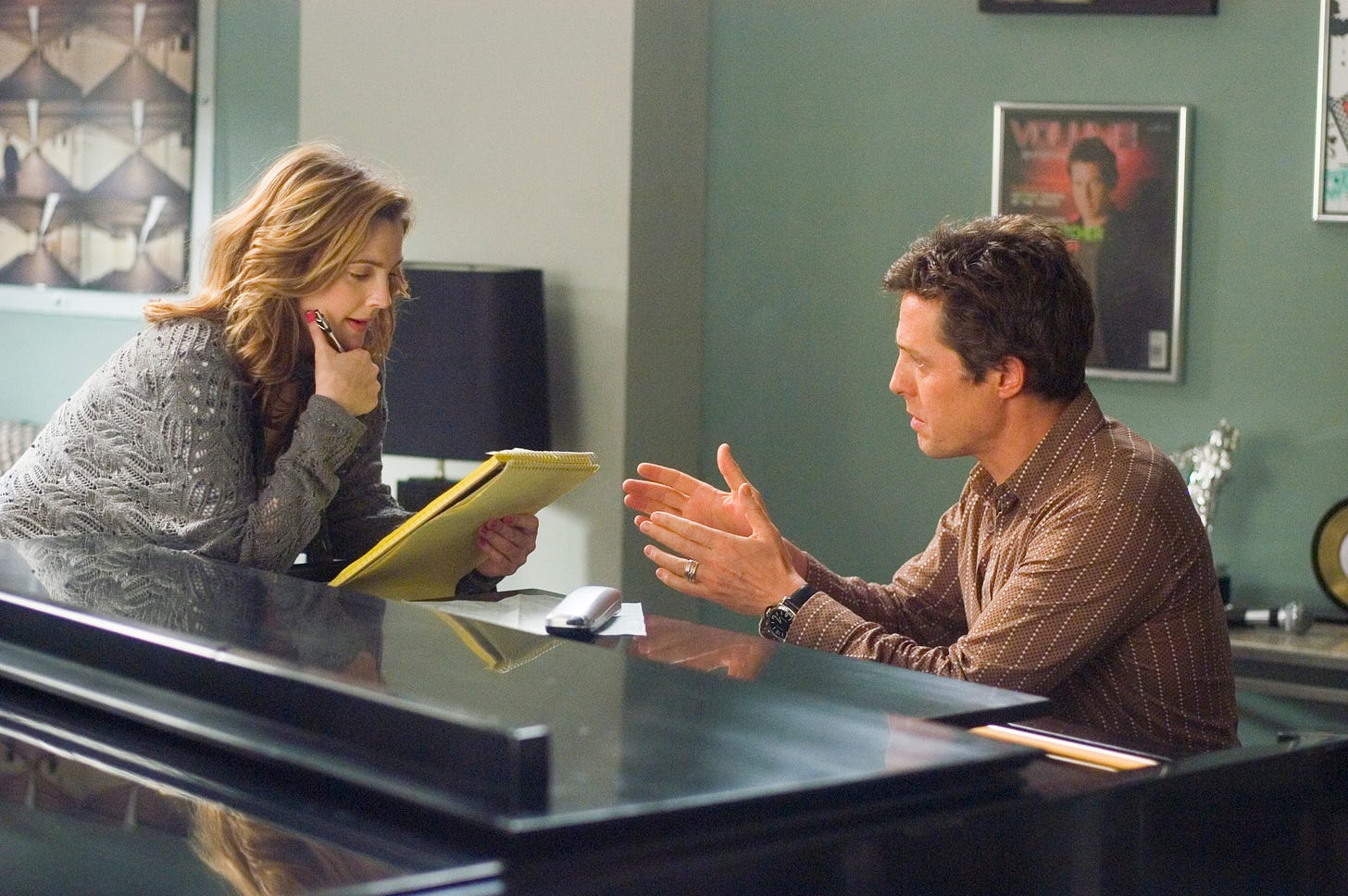 Screencap from Music & Lyrics with Drew Barrymore holding a pen and yellow legal pad and Hugh Grant seated at a piano, talking with her.