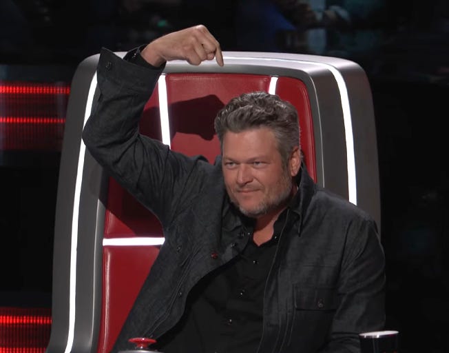 Picture of Blake Shelton from The Voice, where he's sitting in his red coach's chair and wearing all black and pointing down at his own head