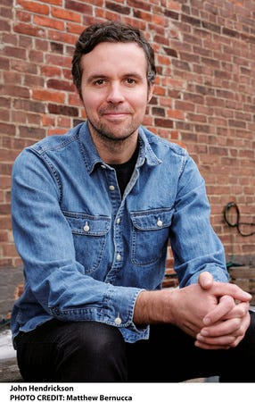 The author, in a denim button-up shirt and black pants, sits in front of a brick wall.