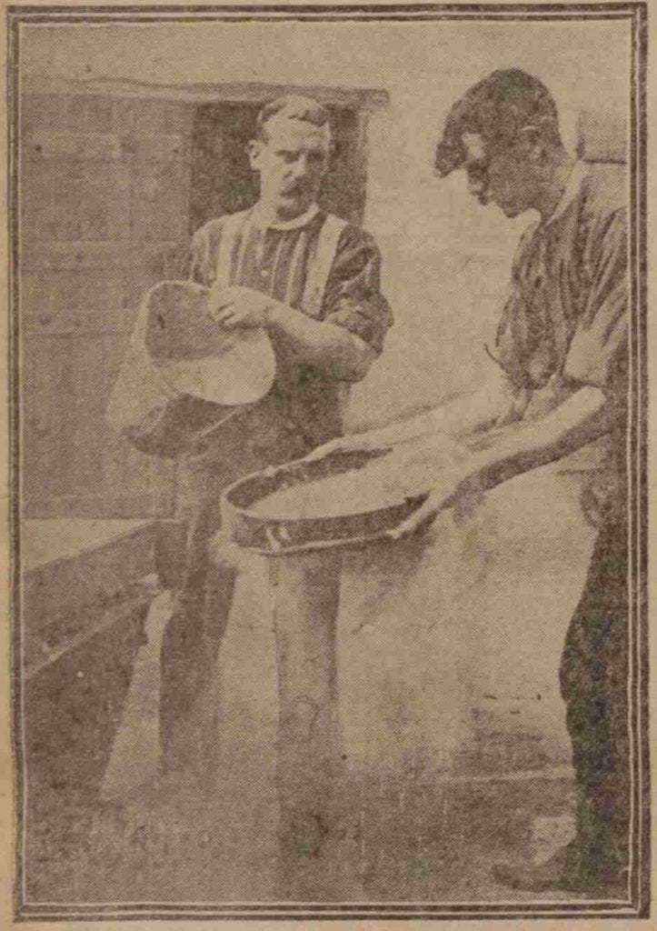 A photograph of two men with moustaches in an outdoor yard in front of a building with an open wooden door. One man holds a large sieve and the other is pouring something from a bucket into it. They both have their sleeves rolled up.