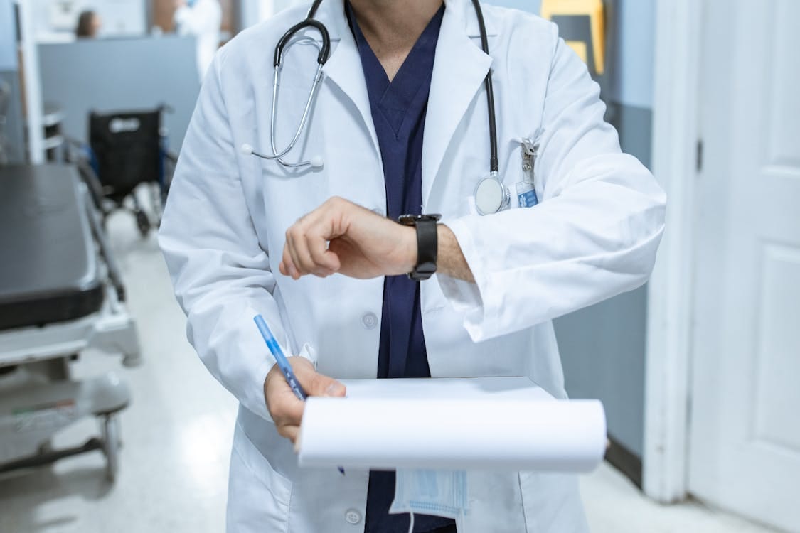 Free Doctor in lab coat checking wristwatch while holding medical chart in hospital corridor. Stock Photo