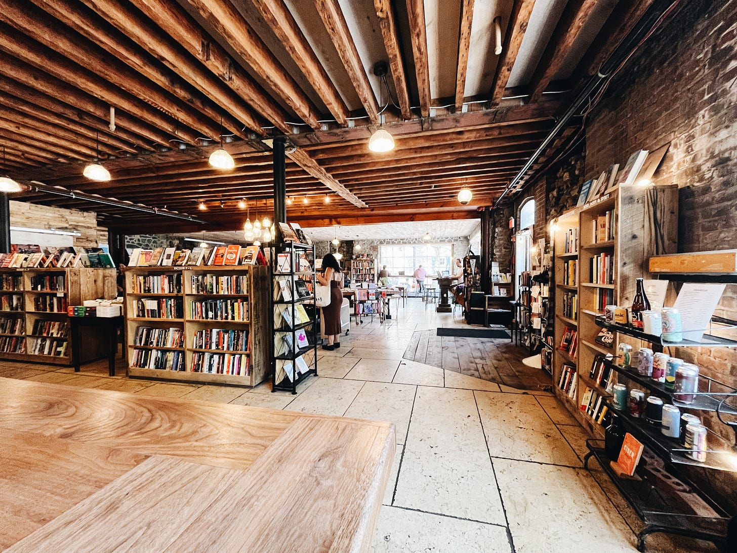 The inside of the beautiful rustic bookstore “Rough Draft” in Kingston, NY with exposed wooden beams on the low ceiling, brick walls, and many bookshelves full of countless colorful books.