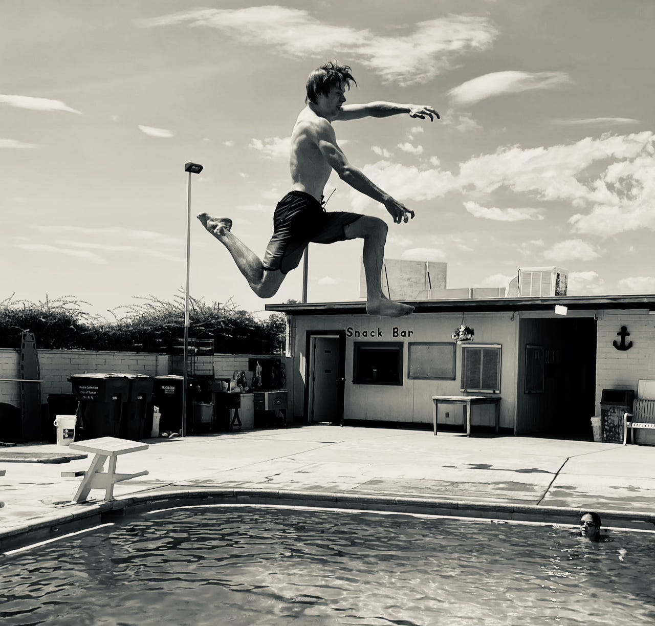 Black and white image of a young man in the air, leaping into a neighborhood swimming pool.