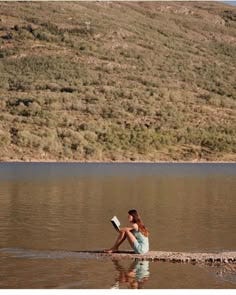 This may contain: a woman sitting on the edge of a body of water with a book in her hand