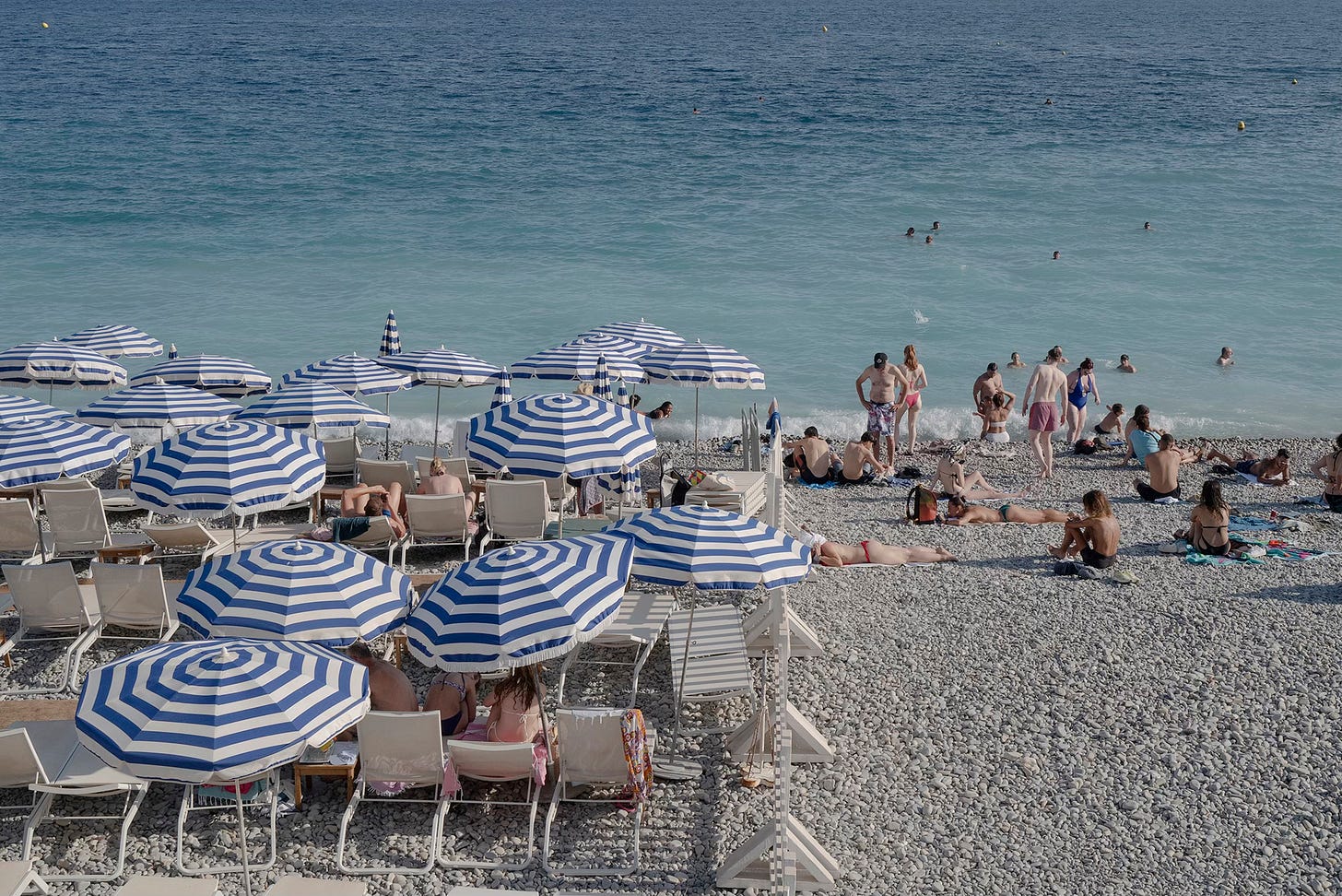 Striped Beach Umbrellas on the Riviera – A picturesque view of Nice’s pebble beach, dotted with neatly arranged blue-and-white striped umbrellas, with sunbathers enjoying the summer weather.