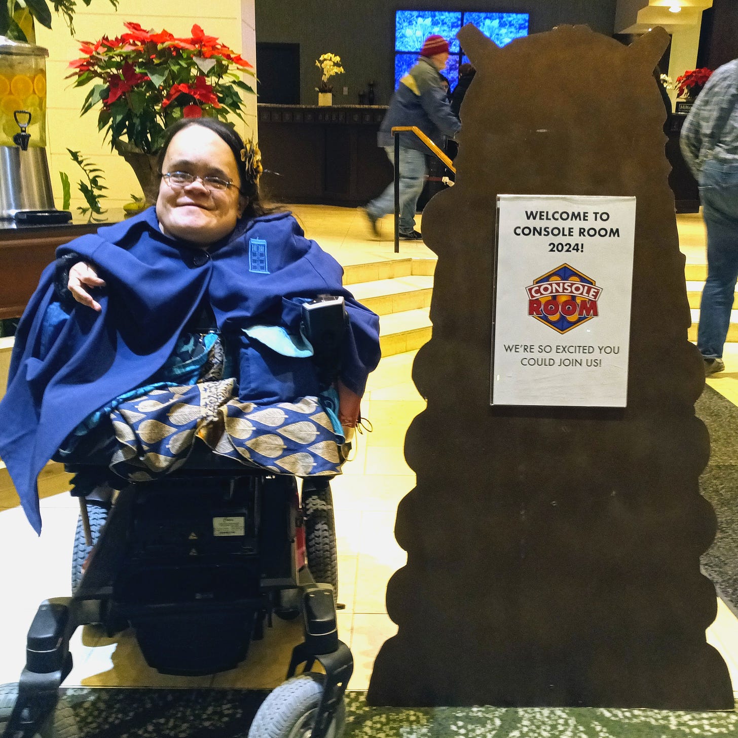 Gaelynn is next to a brass cutout of a Dalek which is posted at the entrance of the Convention with a welcome sign posted on the front. She is smiling and wearing a Doctor Who-themed cloak. It is dark blue and has a TARDIS emblem on the front (the TARDIS is a time machine disguised as a police box). 