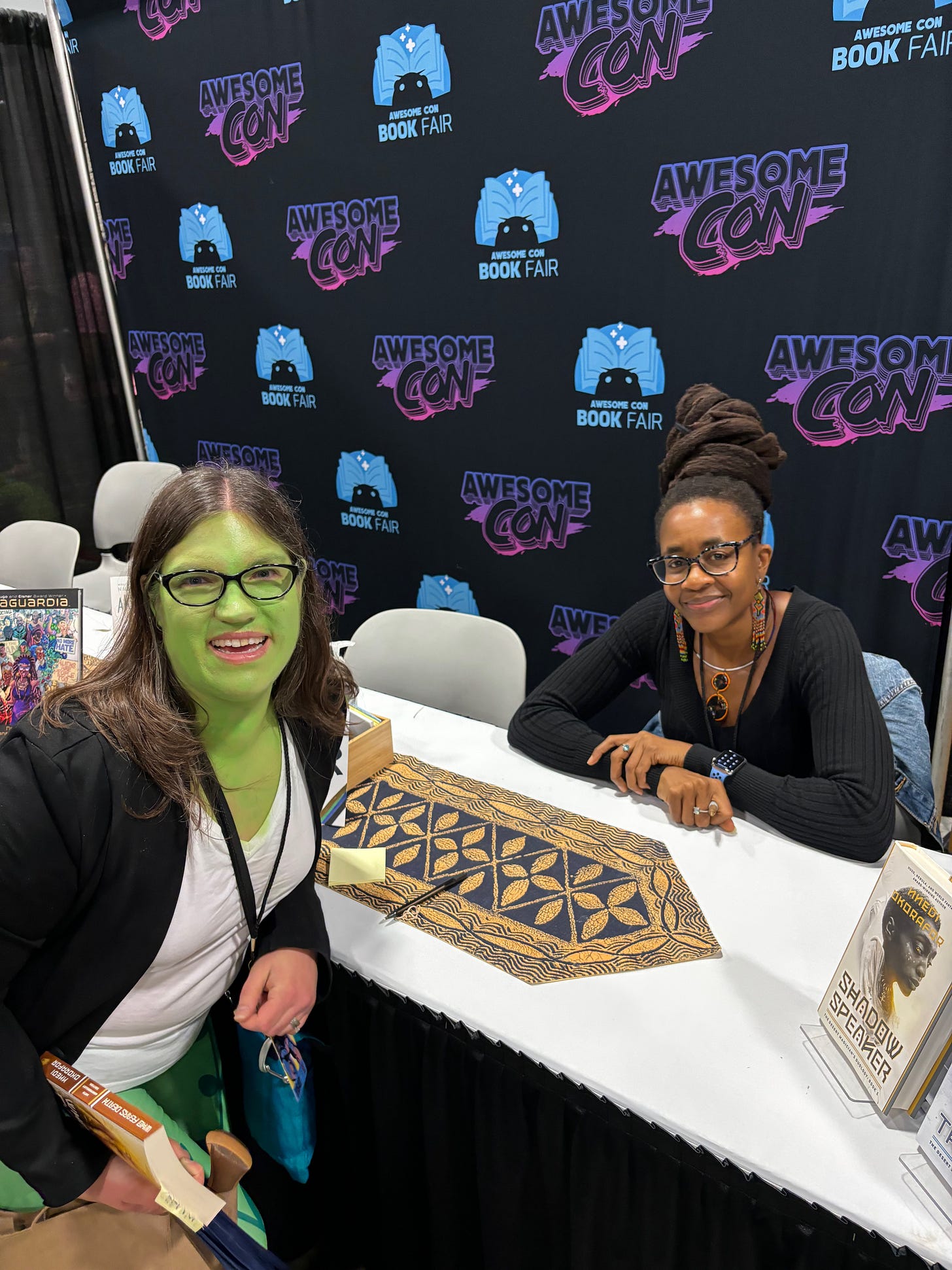 Me (a white woman with a lot of green makeup and a black blazer) crouching down next to a table behind which Nnedi Okorafor (a Black woman in a black sweater) 