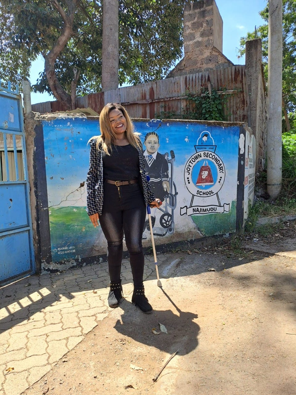 Catherine stands with her cane in front of a blue mural in front of the school