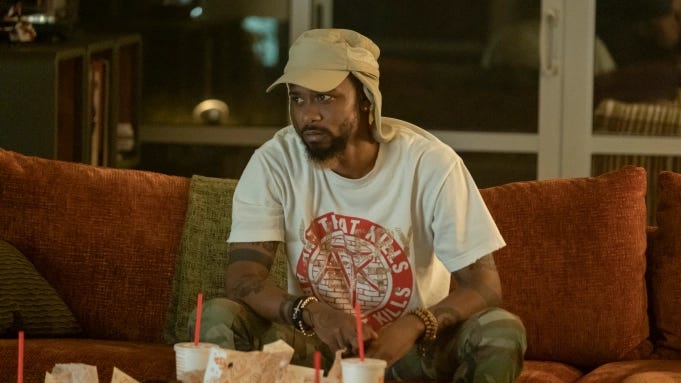 LaKeith Stanfield as Darius, in a light brown cap with neck cover, sitting on a couch in front of takeaways looking intently at an off-screen television in the series finale of Atlanta.