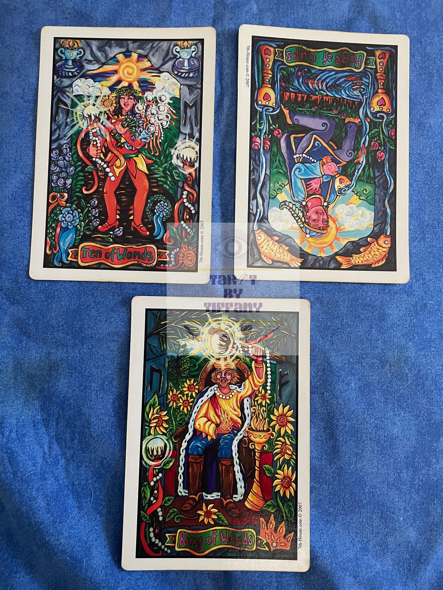 3-card reading using the Pearls of Wisdom Tarot. The cards are laid out on a blue cloth background.Top row (L-R): 10 of Wands, Page of Cups reversed. Bottom card: King of Wands.