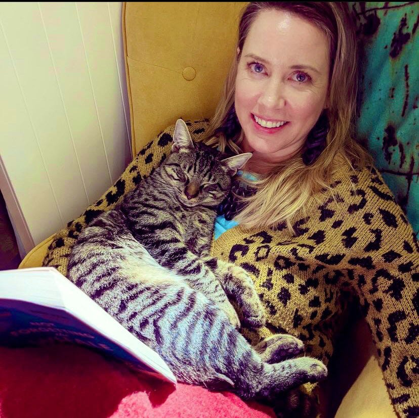 A black-striped, gray cat snuggles up to his person who is propped against a cushion, covered in a red throw and reading a book. She is smiling