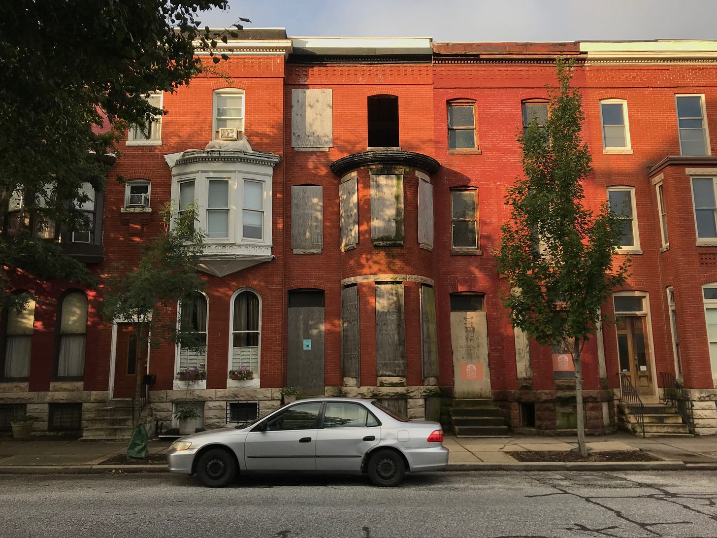 A photograph of red brick three-storey rowhouses, taken from the street. The house in the center has its door and windows boarded.