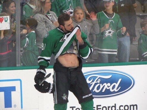 a picture of hockey player tyler seguin from the dallas stars before a game. He is raising the hem of his shirt to wipe his face, displaying his abs in the process. Behind him, a little girl seated in the first row is staring, open-mouthed. 