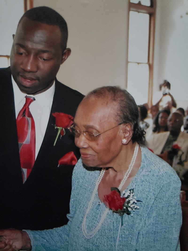 Dark brown skinned man in black suit with a red tie and red rose lapel flower beside older light brown woman with a blue dress and pearls