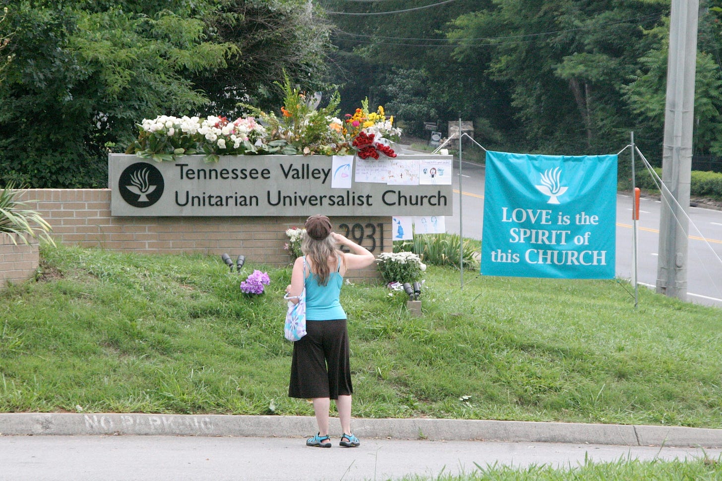 Tennnessee Valley Unitarian Universalist Church sign with many flower bouquets on top, a woman gazing at the sign, and a banner to the side which reads, Love is the Spirit of this Church. 