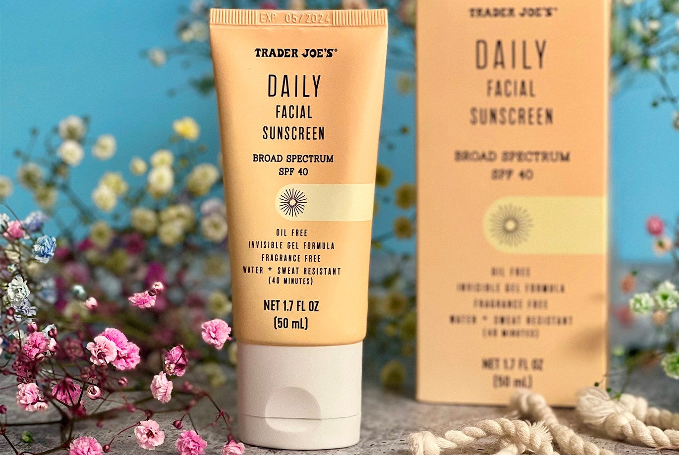 A tube of Trader Joe's Daily Facial Sunscreen surrounded by multicolored flowers, a beach towel, and the box that it comes in.