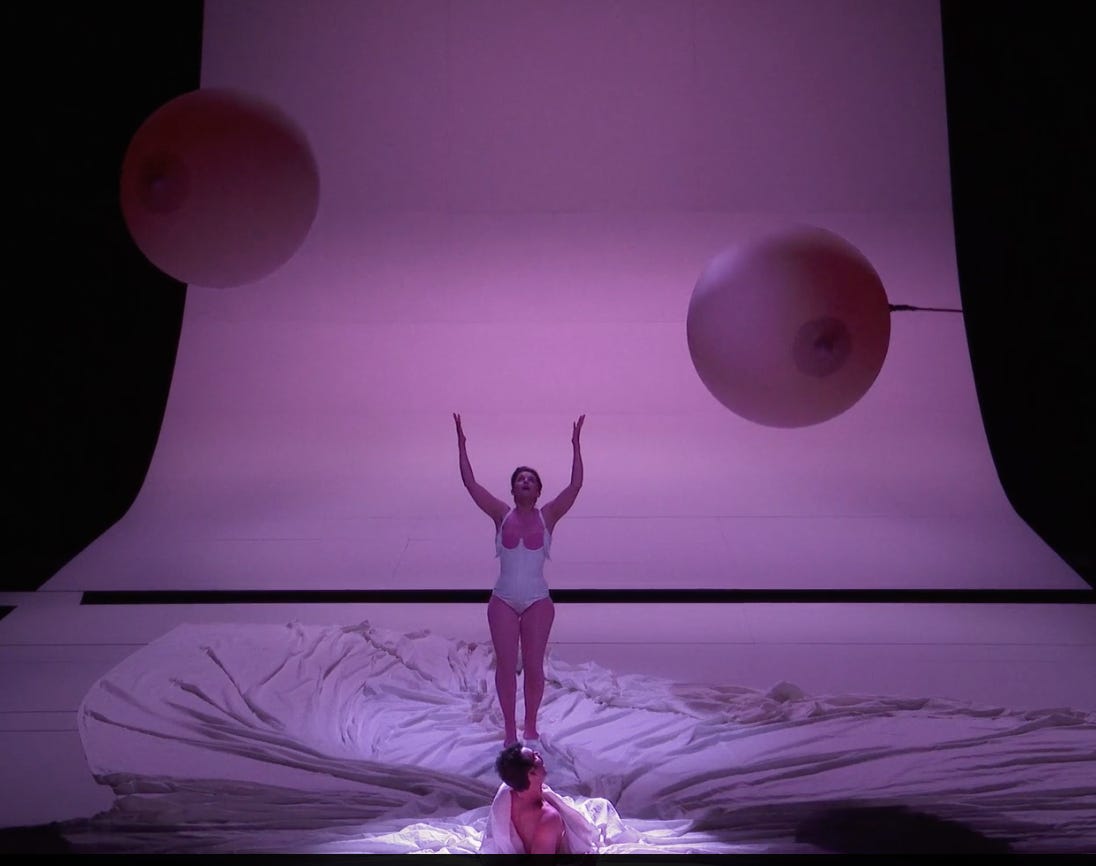 A woman wearing a white corset raises her arms towards two giant balloons shaped like breasts.