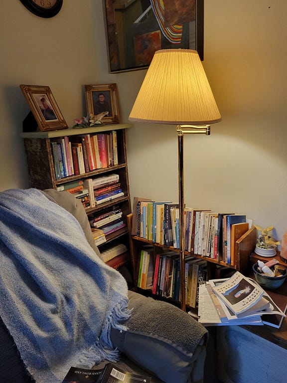 photo of a grey recliner draped with a knitted throw beside a reading lamp and two bookshelves. A clock and painting are on the wall.