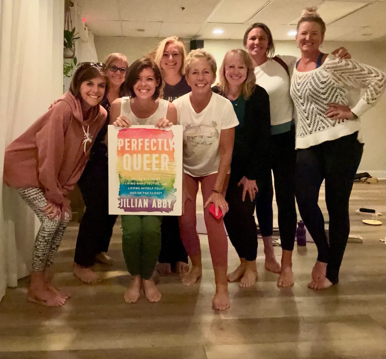 A group of women huddled together and smiling. One is holding a large copy of the book, Perfectly Queer.