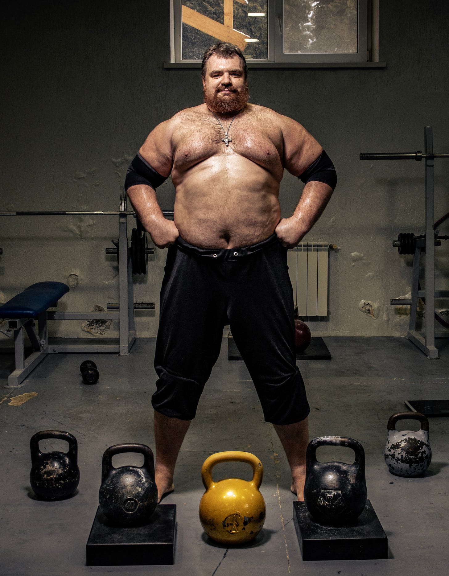 A photo of a strongman standing in a gym surrounded by kettlebells.