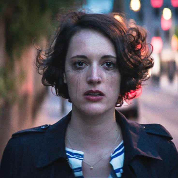 A woman with short, curly dark hair walks down a city street at dusk, her face streaked with tears. She wears a dark coat over a striped shirt, looking distressed.