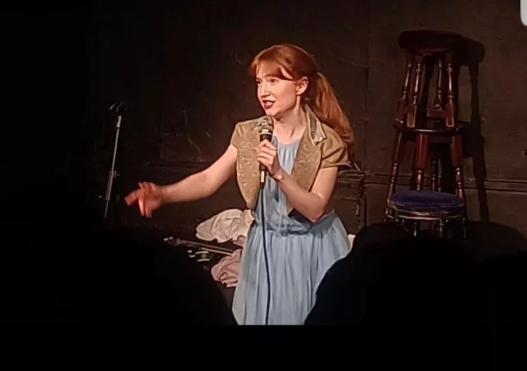 Small ginger haired woman on stage talking into a microphone wearing a light blue dress and small gold jacket