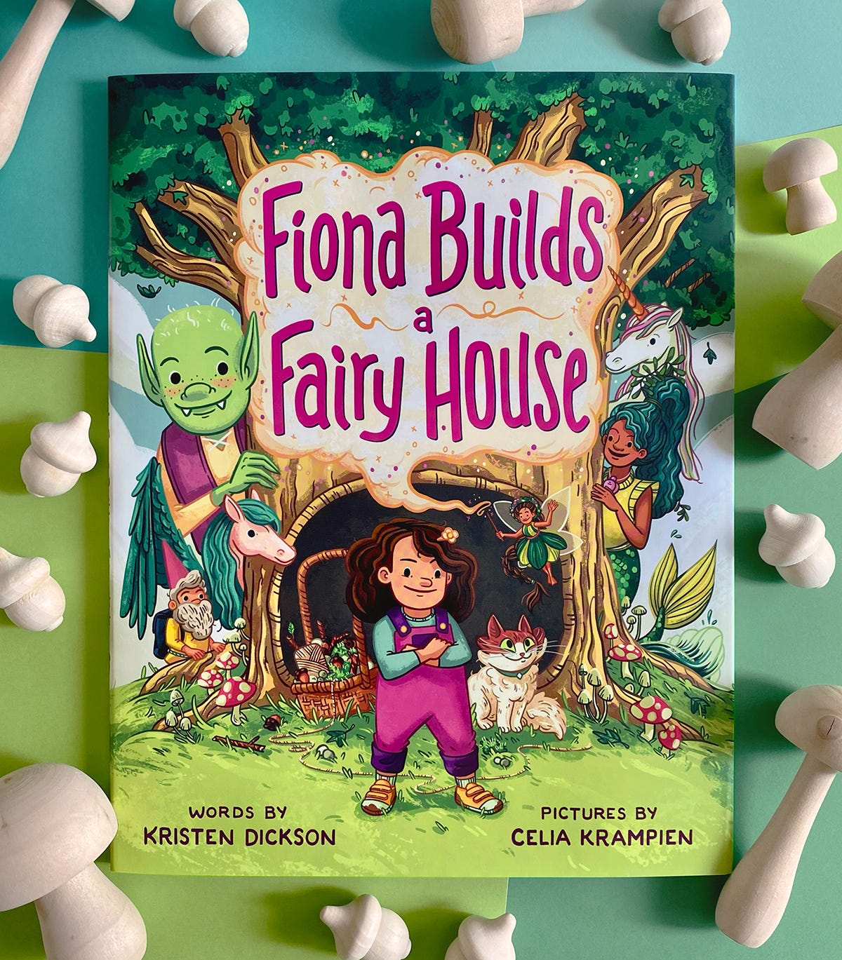 Book FIONA BUILDS A FAIRY HOUSE sitting on a bright paper background surrounded by wooden mushrooms and acorns. The cover shows a young girl with dark, curly hair and bright pink overalls standing in front of a huge tree with a round hollow. She's surrounded by smiling fairytale creatures as well as a cat and basket of foraged building supplies.