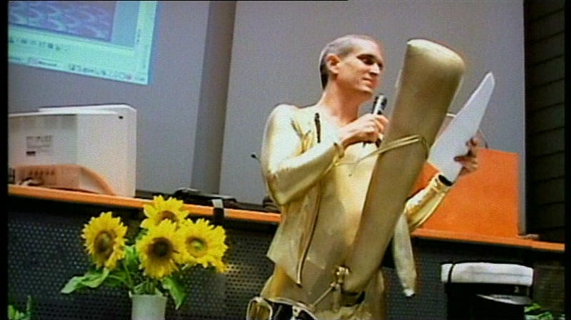 A man stands in a gold suit with an inflatable penis reading off of a script.