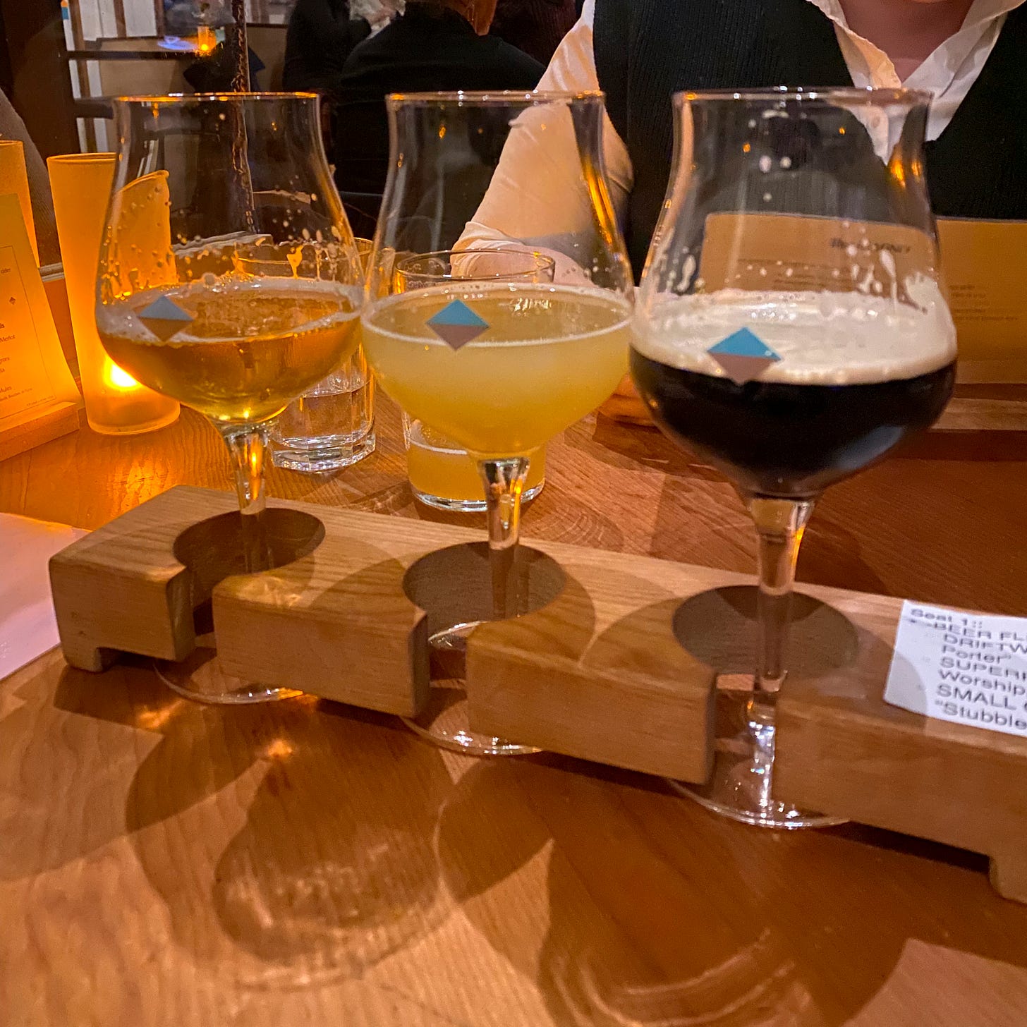 Three tulip glasses of different beers in a wooden flight paddle on a wood table. A candle and a glass of water are behind them and to the left. Across the table is someone in a sweater vest and a white shirt.