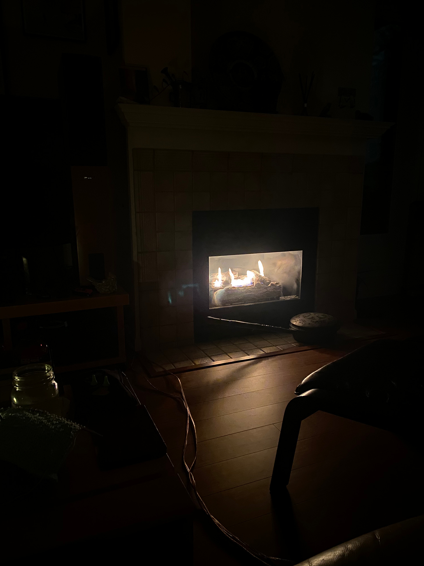 A dark image of the living room, the only light from the fireplace in the back centre. A few shapes are visible in the foreground: an ottoman, the coffee table with a glass of water on it.