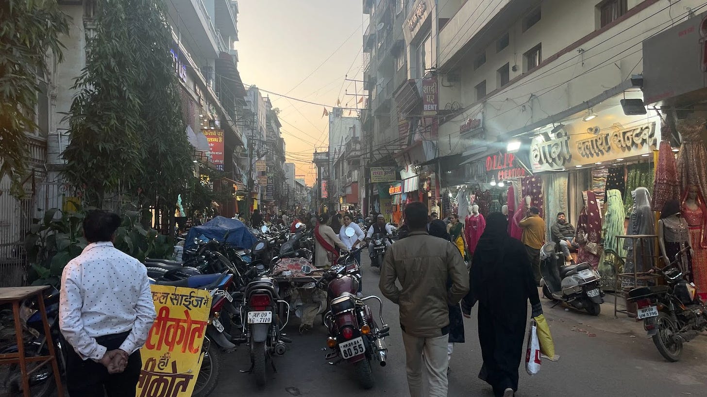 Crowded Indian street. Parked motorcycles take up much of the road. People walk and there are sari shops lining the sides.