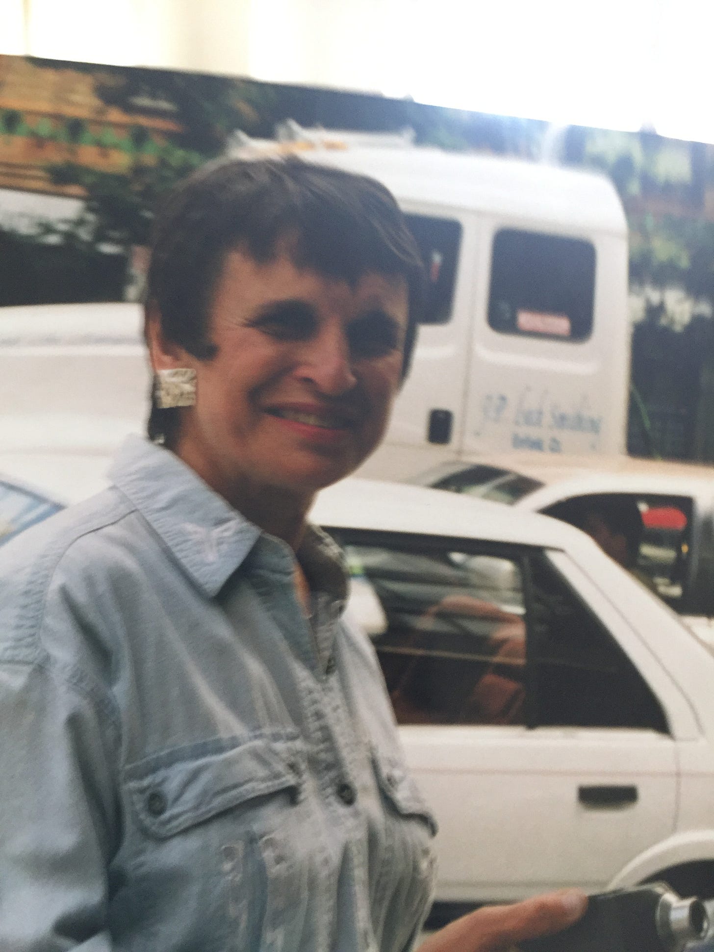 Dr. Arline Bronzaft, an older woman with short hair, smiling on a city street as she holds an old-fashioned decibel reader. Behind Dr. Bronzaft, an environmental psychologist and expert on noise, there's a small sports car and behind that a large semi-truck in the street.