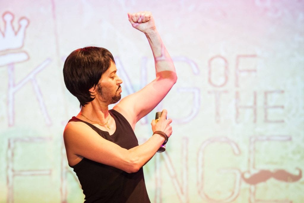 masc presenting white person in vest and briefs sprays deo into armpit on stage.