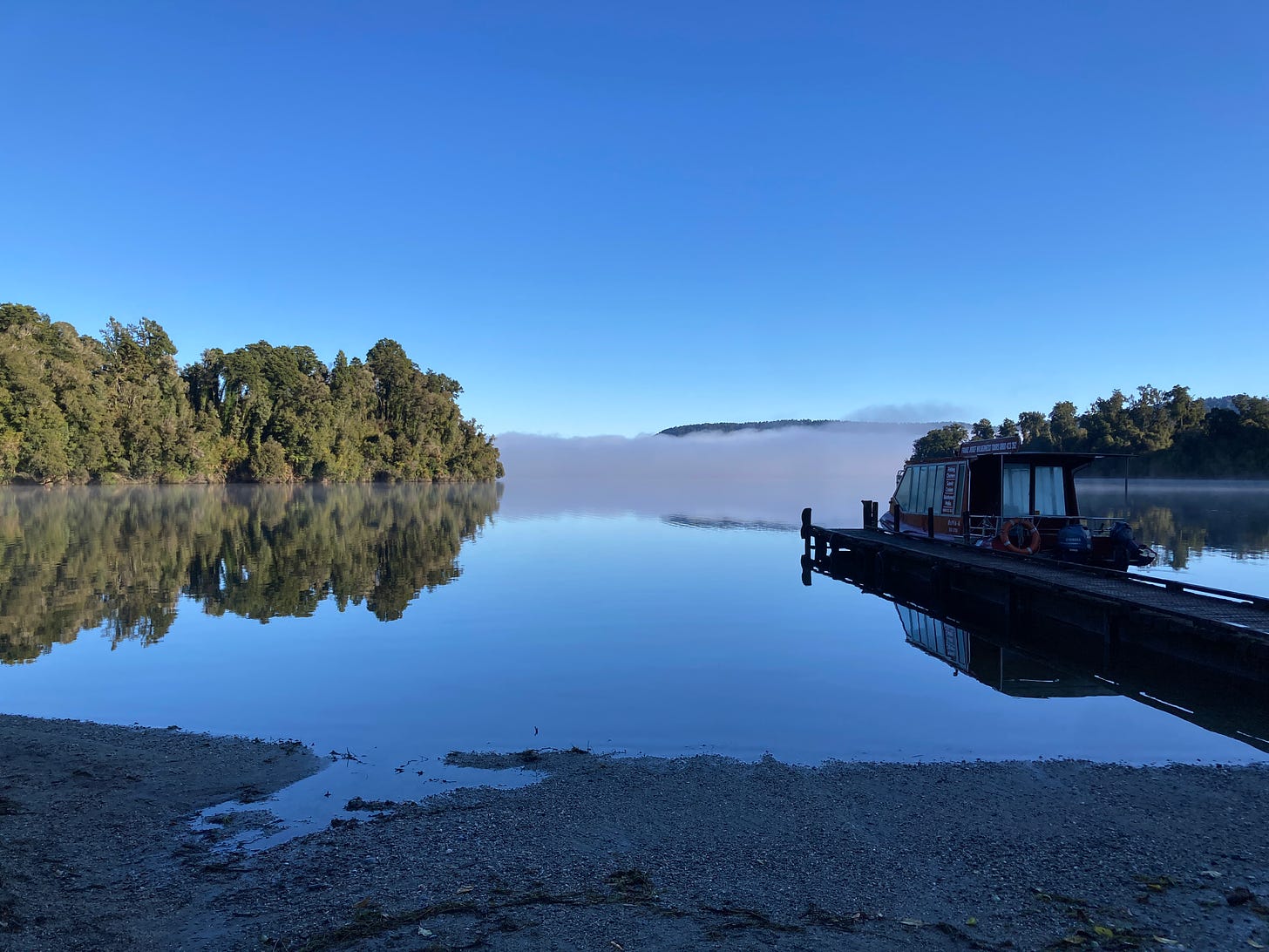A picture of a lake in the morning, the landscape perfectly reflected in the water. On the right, a lone boat waits tied up a pier jutting out in to the water. In the near distance, mist blankets the view beyond.