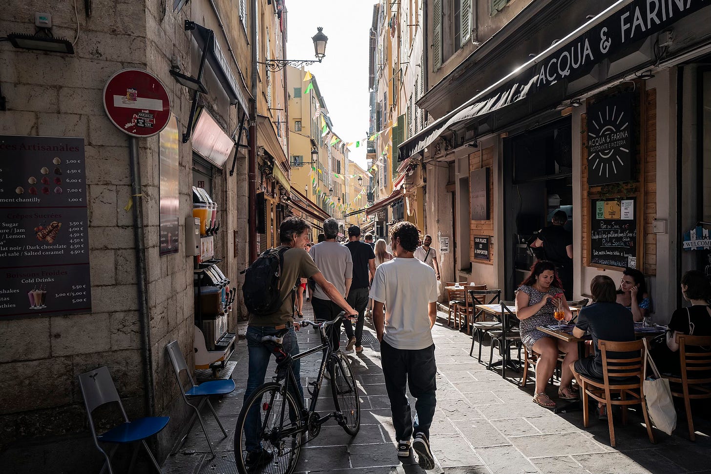 A sunlit cobblestone street in Nice, France, where people are walking past a small café with outdoor seating. A cyclist and a pedestrian are engaged in conversation.