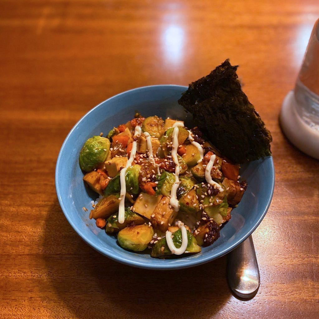A blue bowl of the stir-fry described above, with pieces of carrot, tofu, and brussels sprouts in sauce under a drizzle of kewpie mayo and a dusting of sesame seeds. At the edge of the bowl are two sheets of gim.