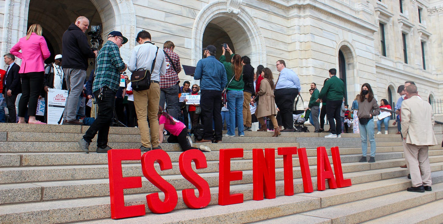 a crowd of people standing on the state capitol steps at a press conference. Red letters spell out the word "essential" in all caps on the steps