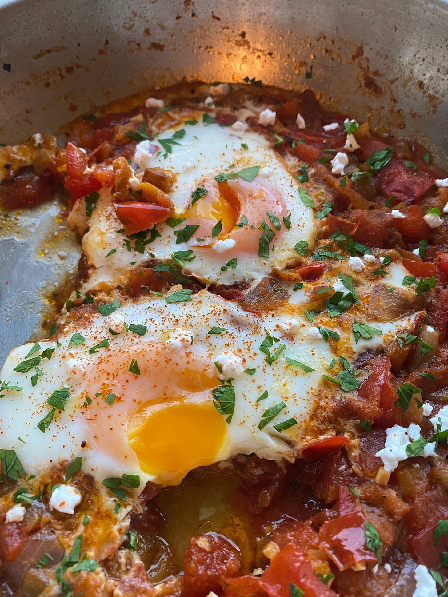 A close-up of a pan of shakshuka, already partially dished out. The egg yolks have been broken open and they're running a little. Everything is topped with parsley and feta.