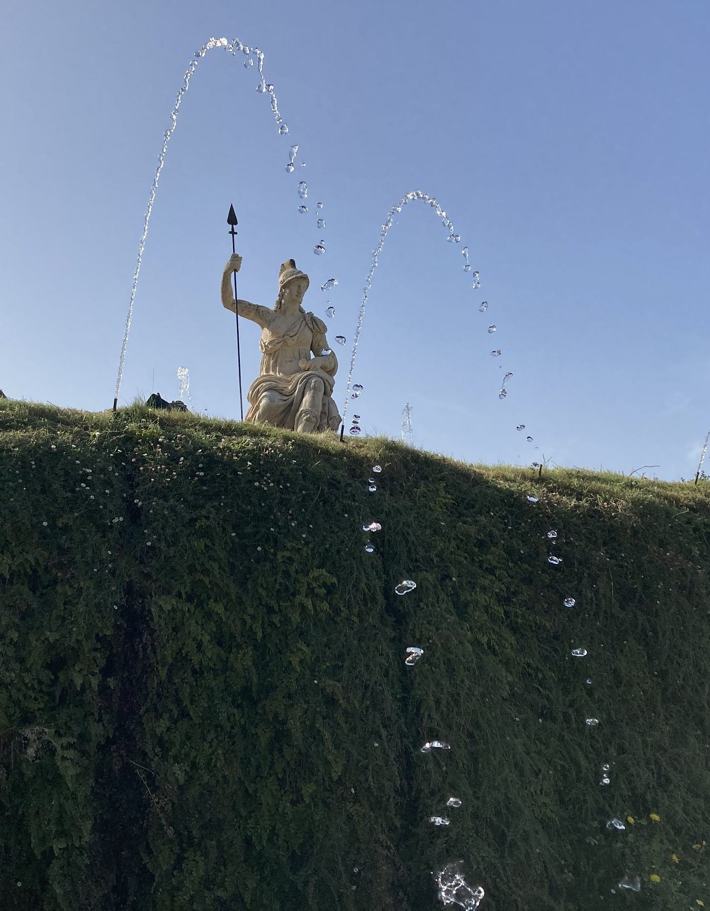 Two arcs of water against blue sky and a fern-covered wall, with a marble statue