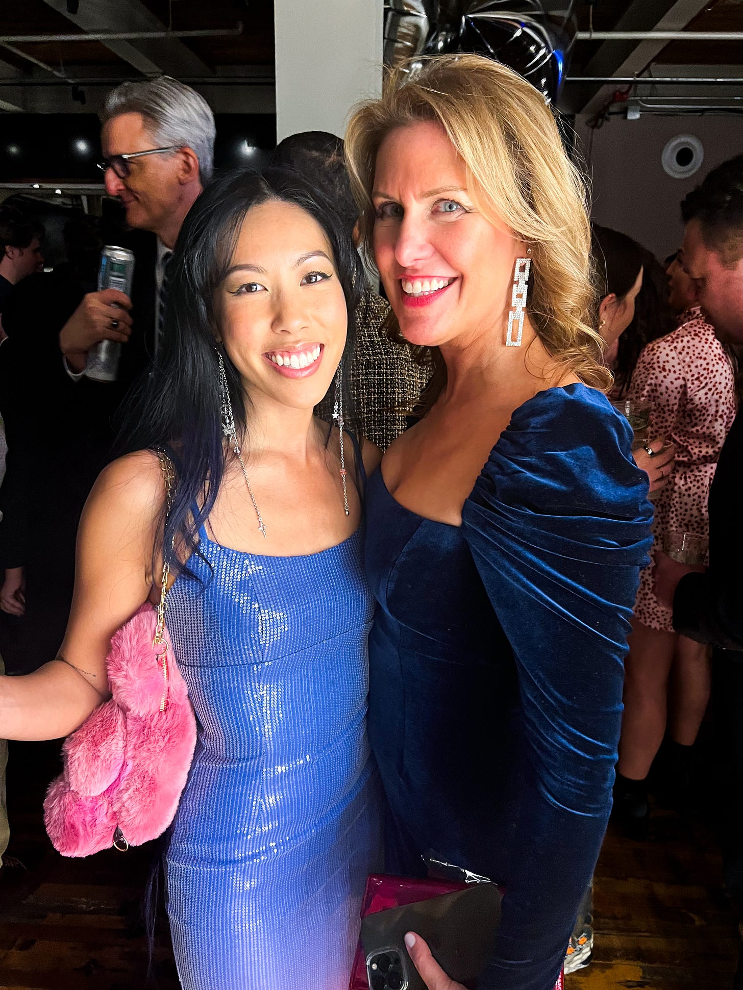 Two women in elegant blue dresses smiling at the camera during a party.