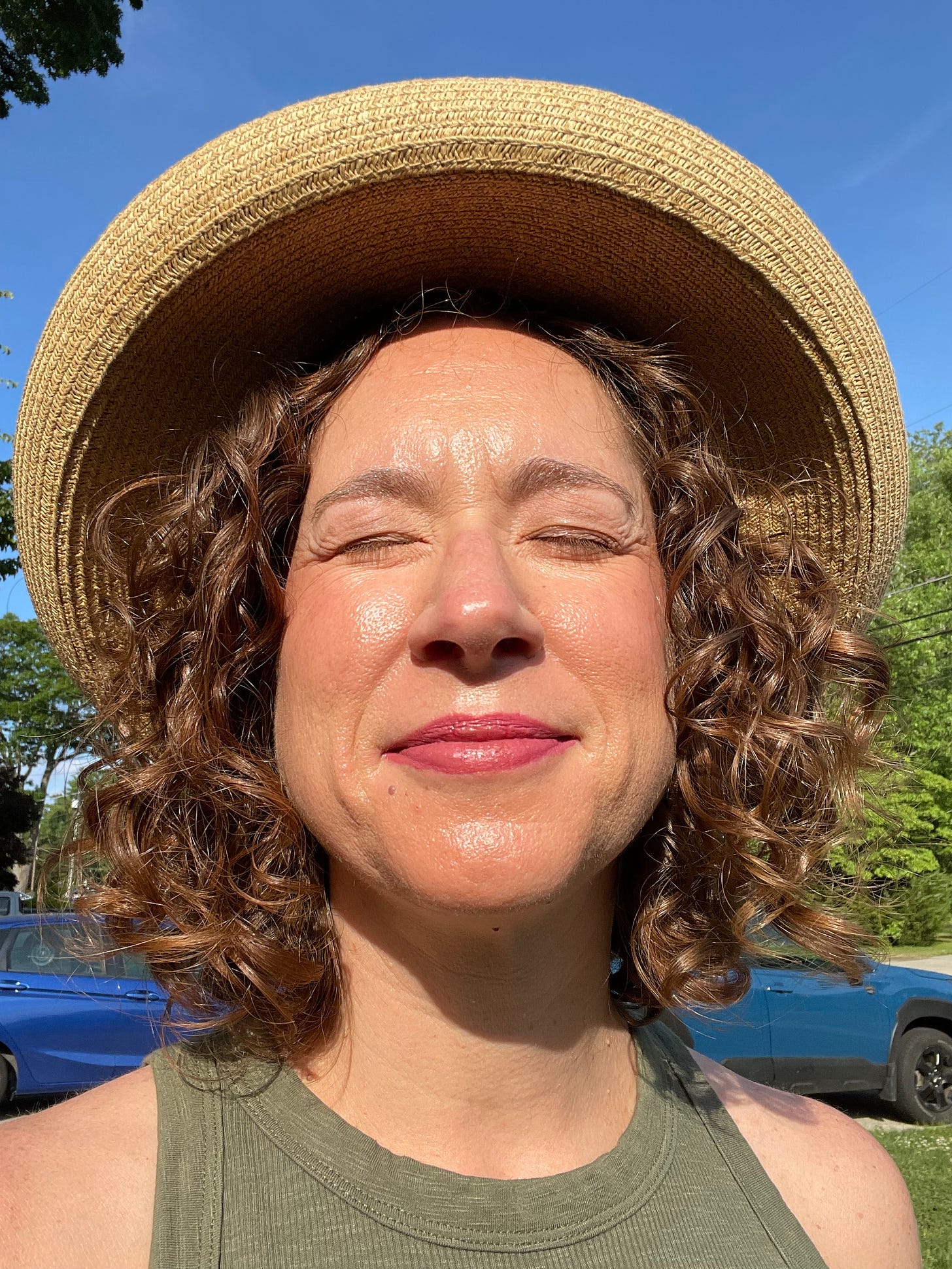 My face in the sun. I'm wearing a straw sun hat and olive green tank top. My eyes are closed and I'm smiling.